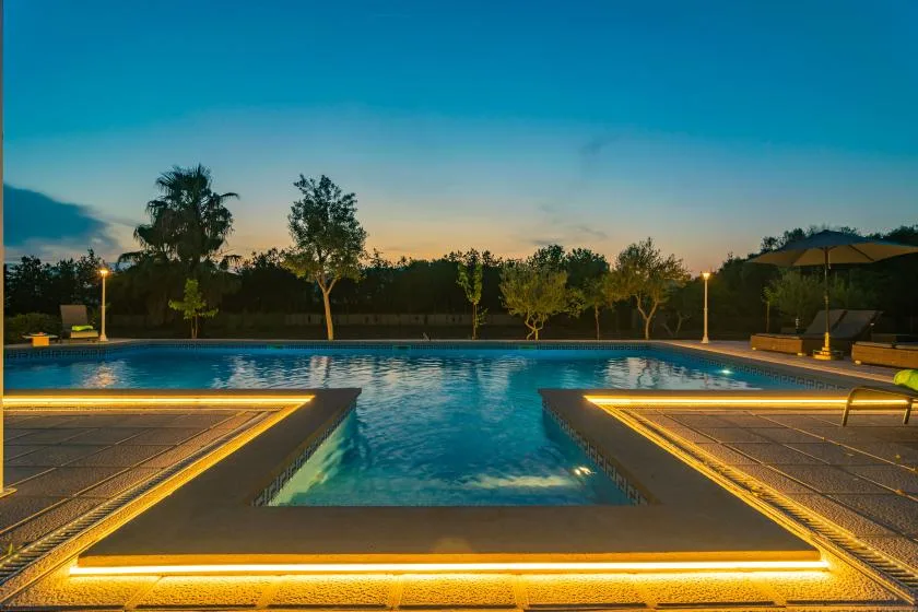An illuminated outdoor swimming pool at dusk, with trees along the far edge.