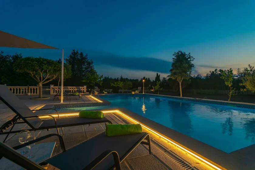 An illuminated outdoor pool sits beside a paved deck with lounge seating at dusk.
