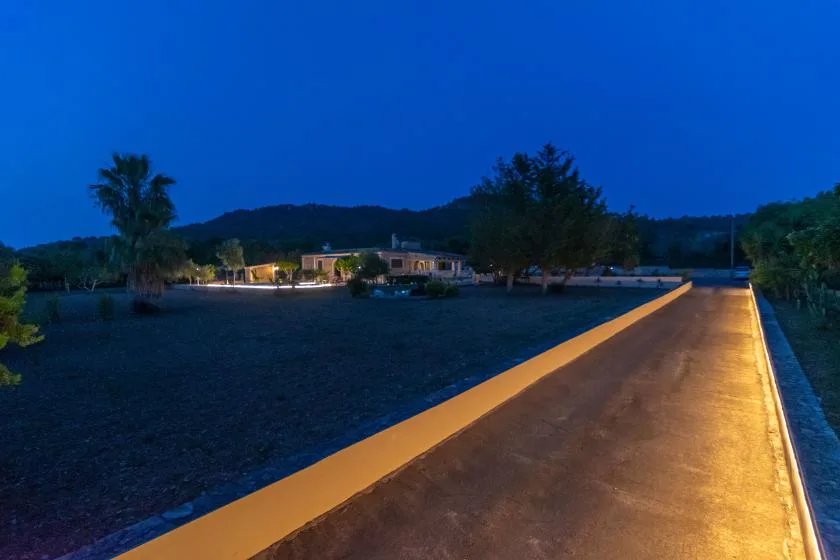 A lit driveway leads toward a low building beneath a deep blue evening sky.
