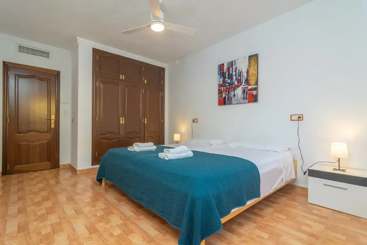 A bedroom with a bed covered by a teal blanket and a wooden wardrobe.