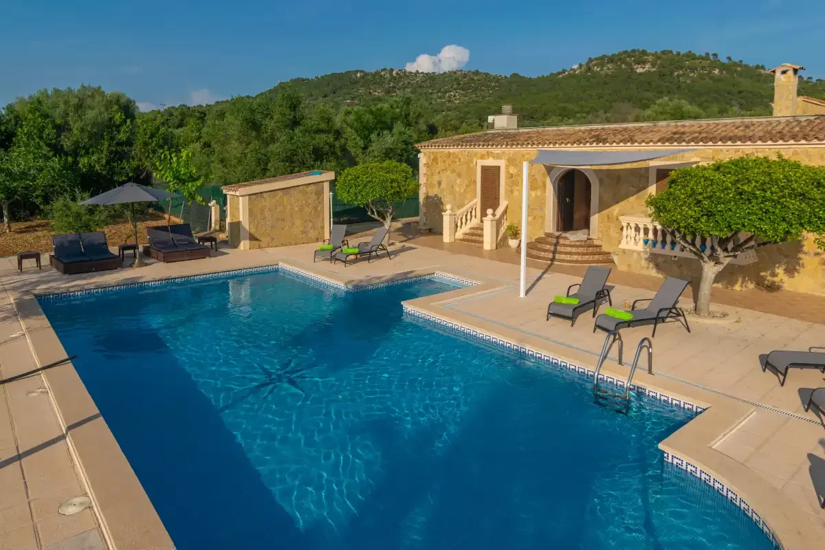 A swimming pool beside a stone-colored house, with loungers on the surrounding patio.
