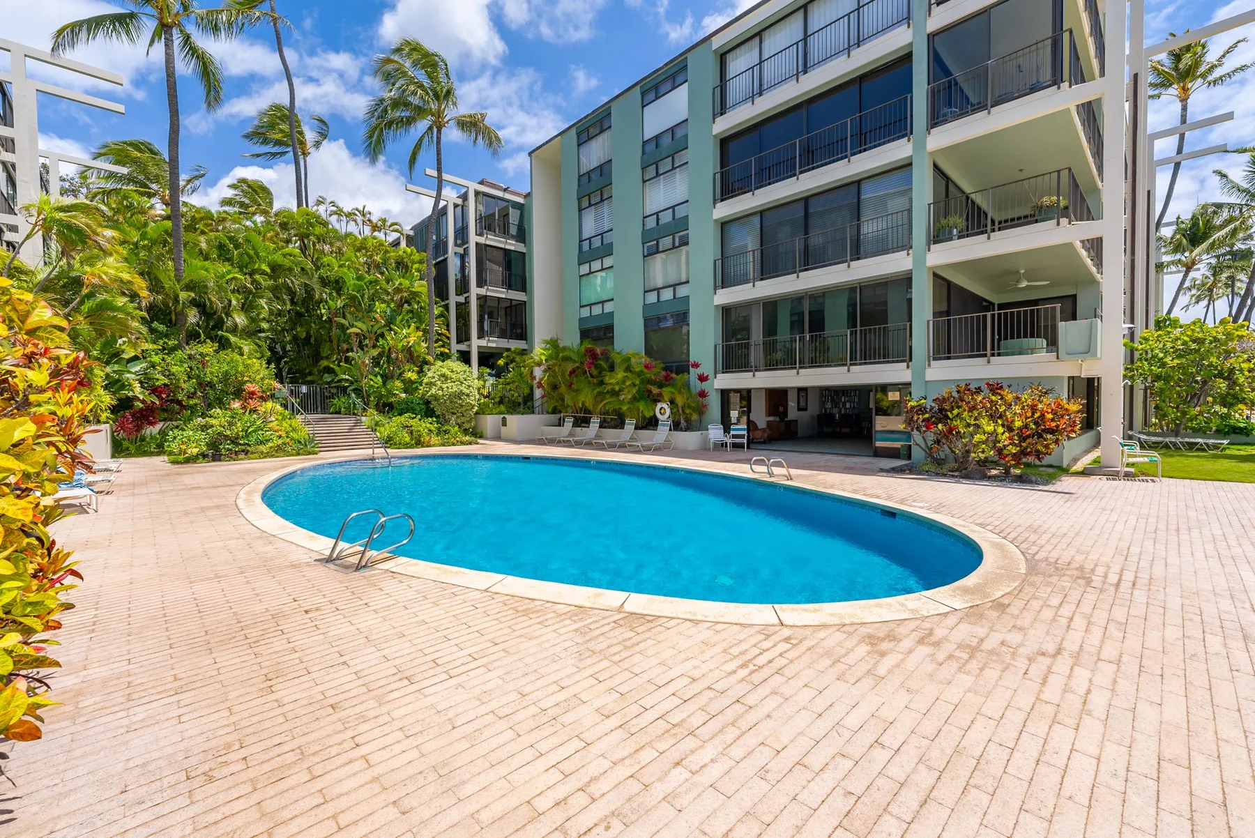 An outdoor swimming pool sits beside a multi-story building and tropical landscaping.