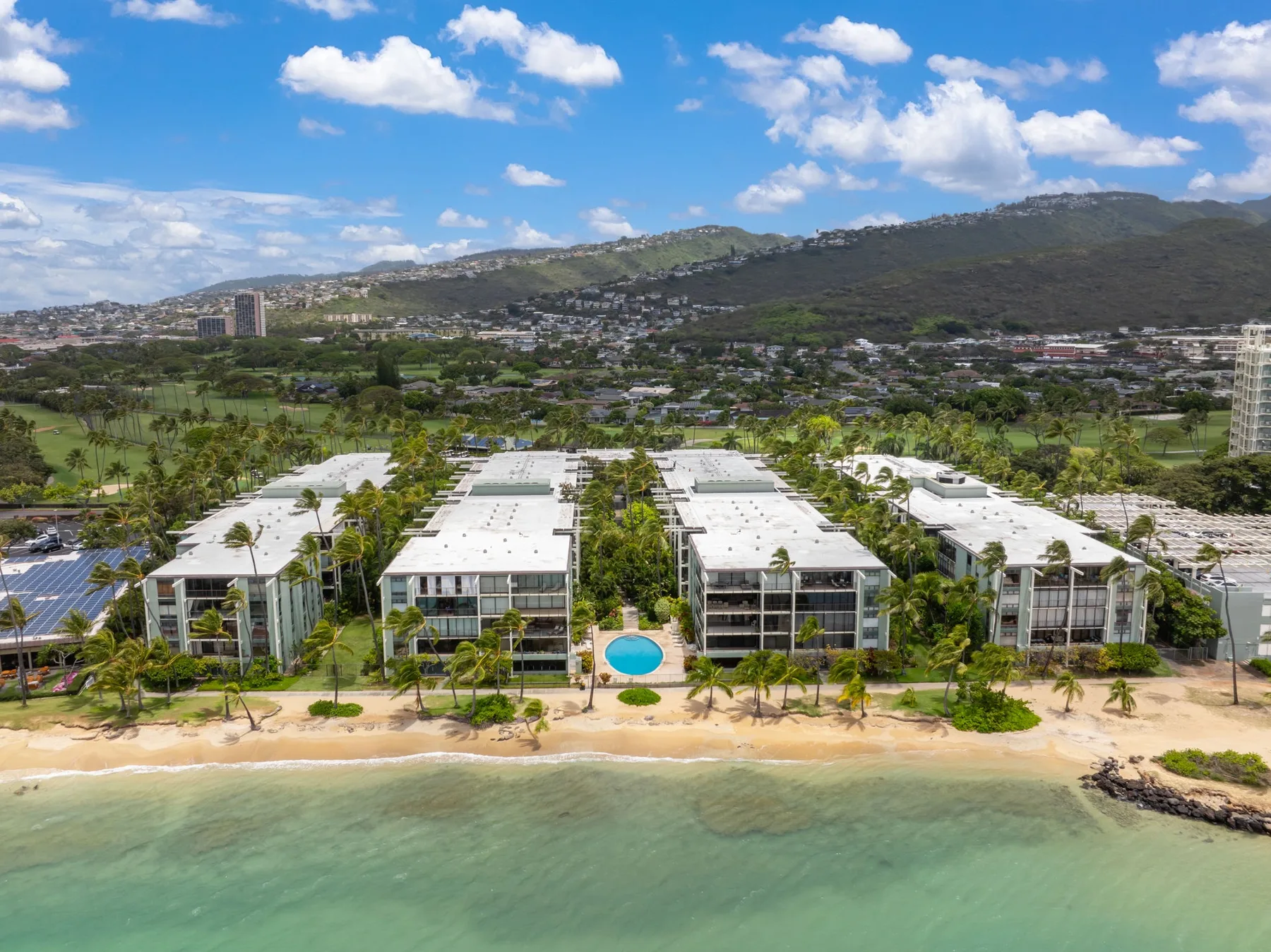 Aerial view of beachfront resort buildings, a sandy shore, and green hills beyond.