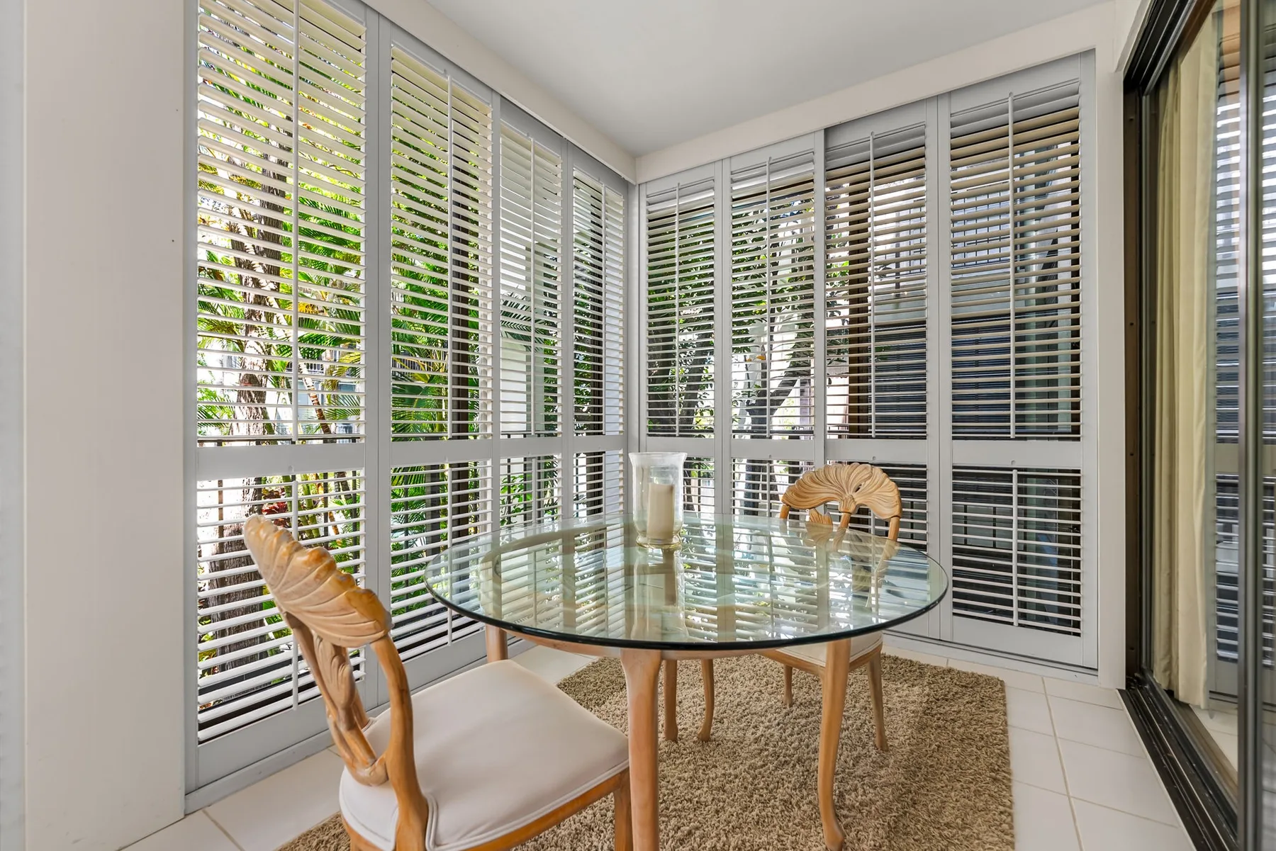 A bright dining nook with a round glass table beside wide windows with blinds.