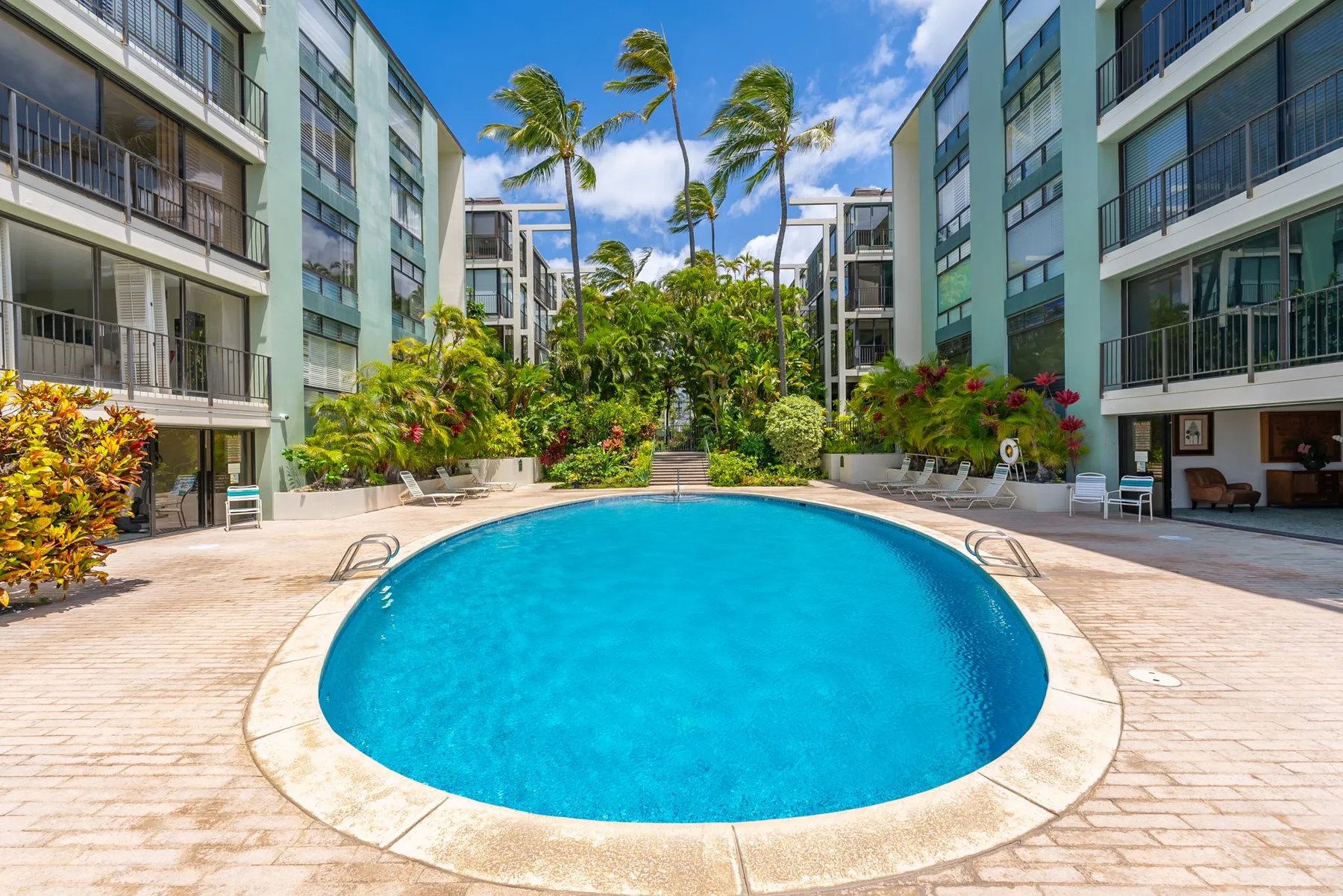 A swimming pool sits between modern buildings, surrounded by tropical plants.