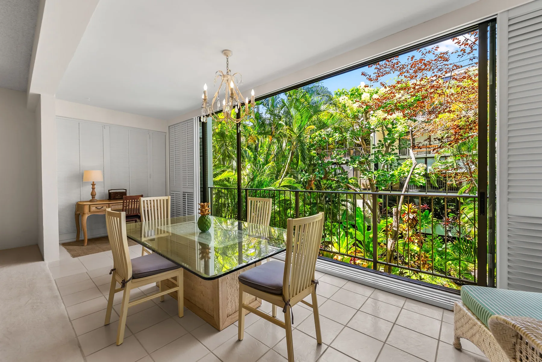 A bright dining area has a glass table beside large windows overlooking greenery.