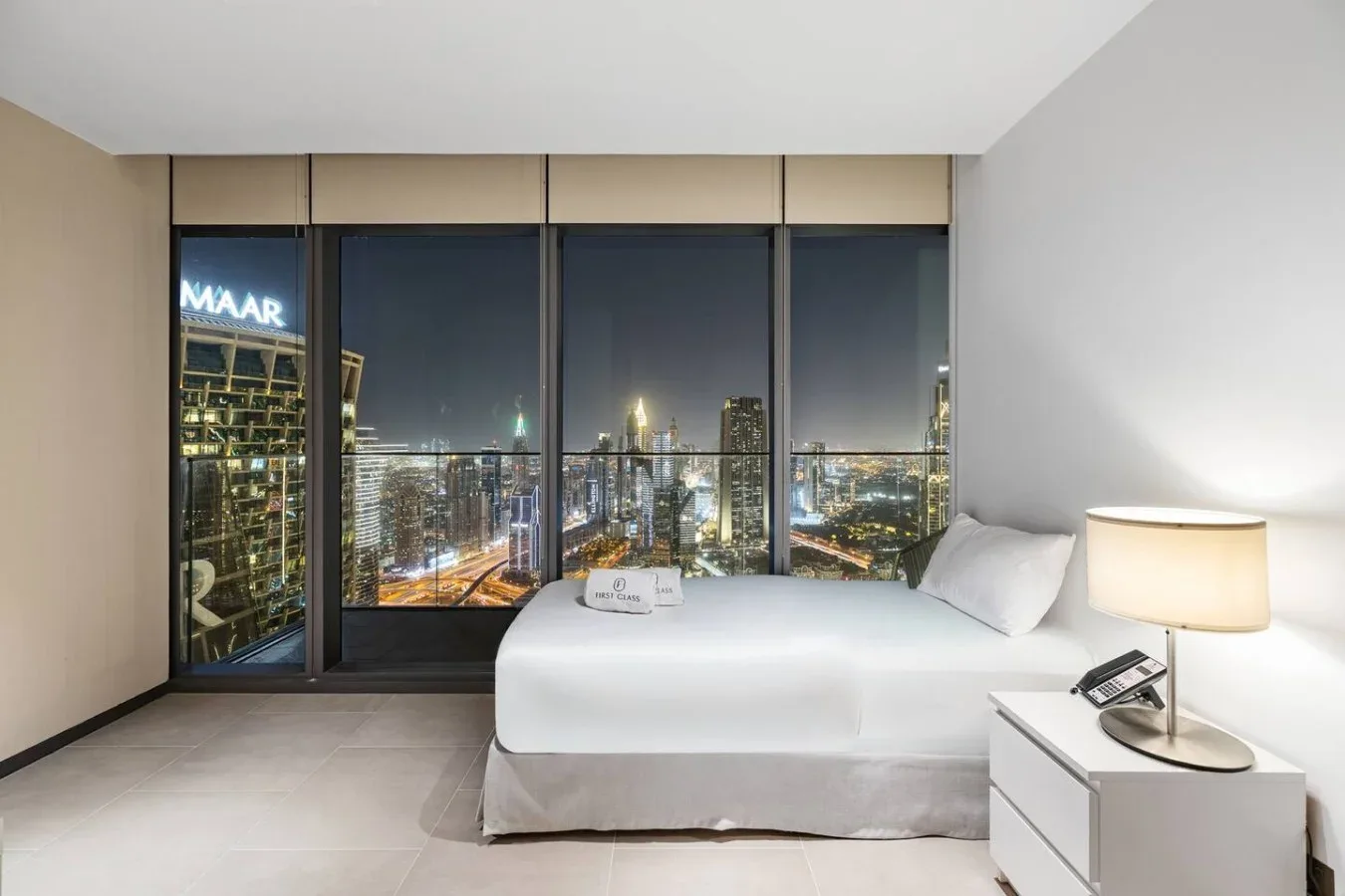 Dormitorio con una cama junto a grandes ventanales con vistas a la ciudad de noche.