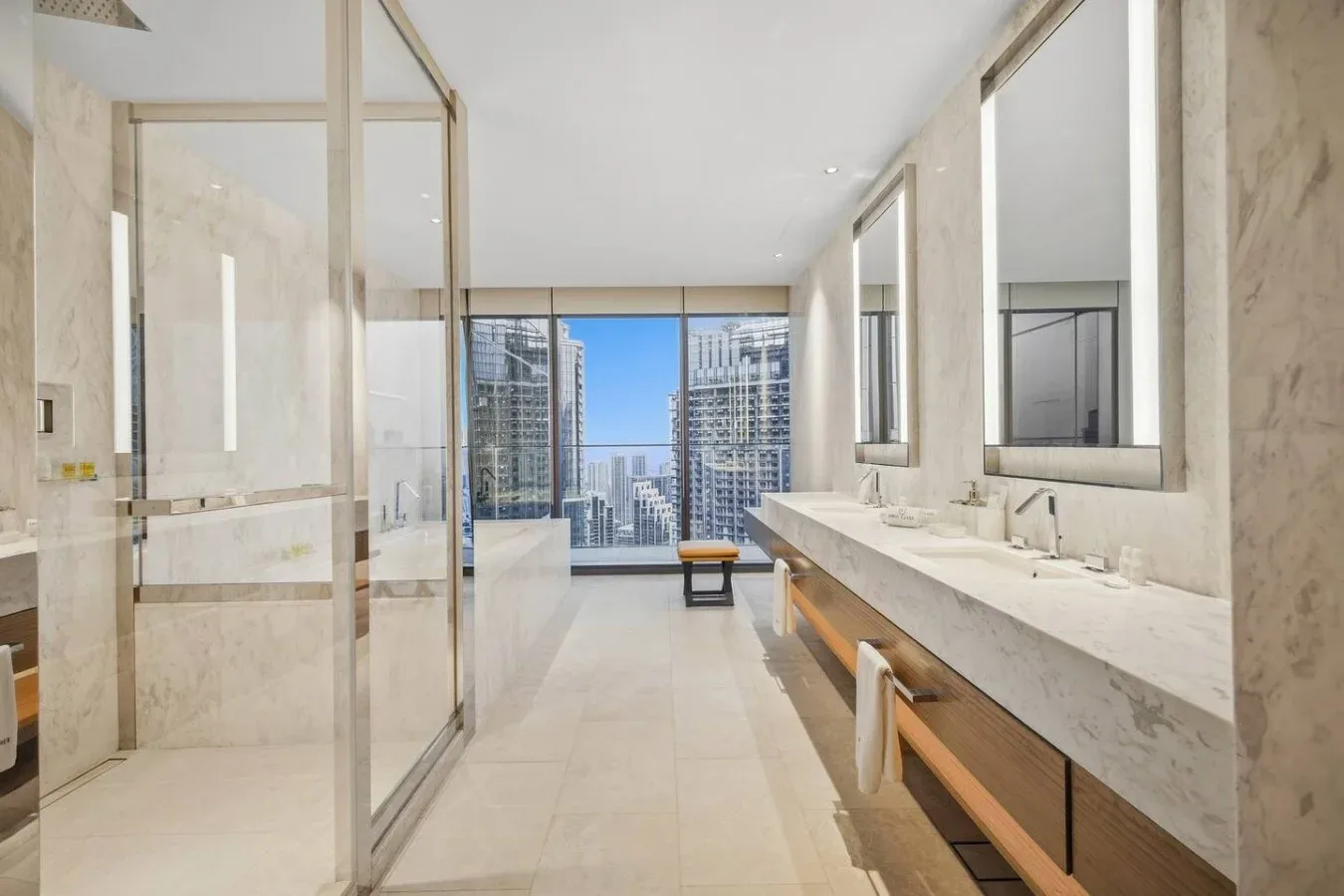 Un baño luminoso con un largo tocador y una ducha de vidrio, con vistas a edificios de la ciudad.