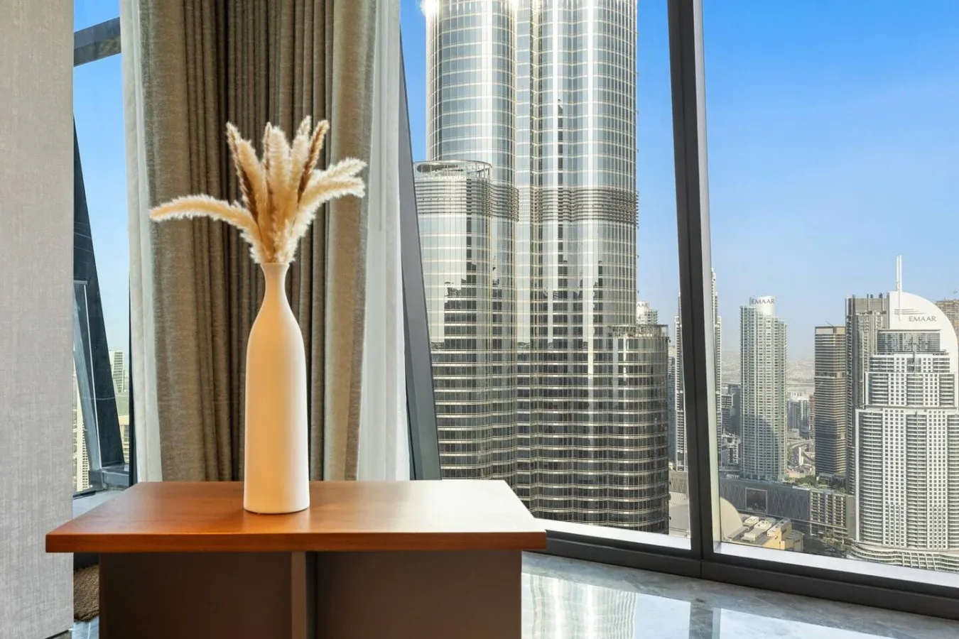 Un jarrón con hierbas secas está sobre una mesa junto a una ventana grande con vistas a edificios.
