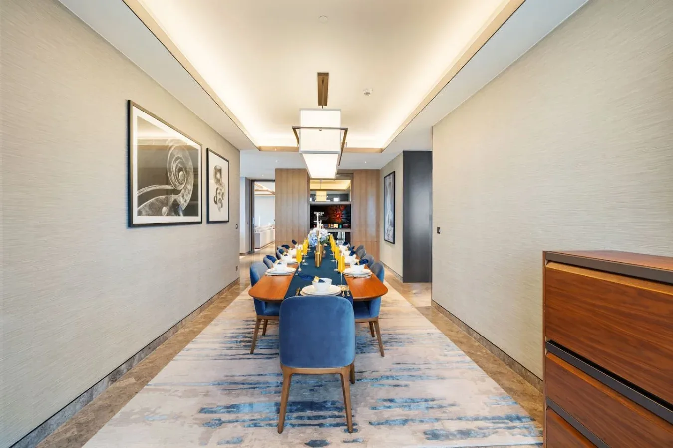 Un comedor alargado tiene una mesa puesta, sillas azules y cuadros en las paredes.