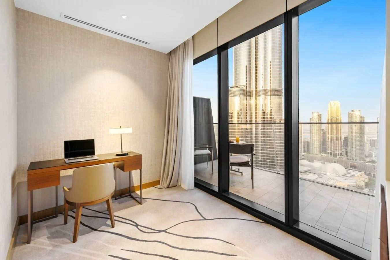 Una habitación luminosa con escritorio y asientos junto a grandes ventanales con vistas a edificios.