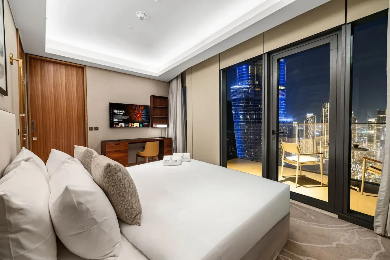Un dormitorio con cama, escritorio y ventanas de piso a techo con vistas a las luces de la ciudad.