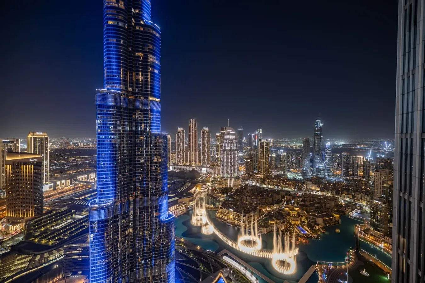 A nighttime city skyline with illuminated skyscrapers and a brightly lit fountain.