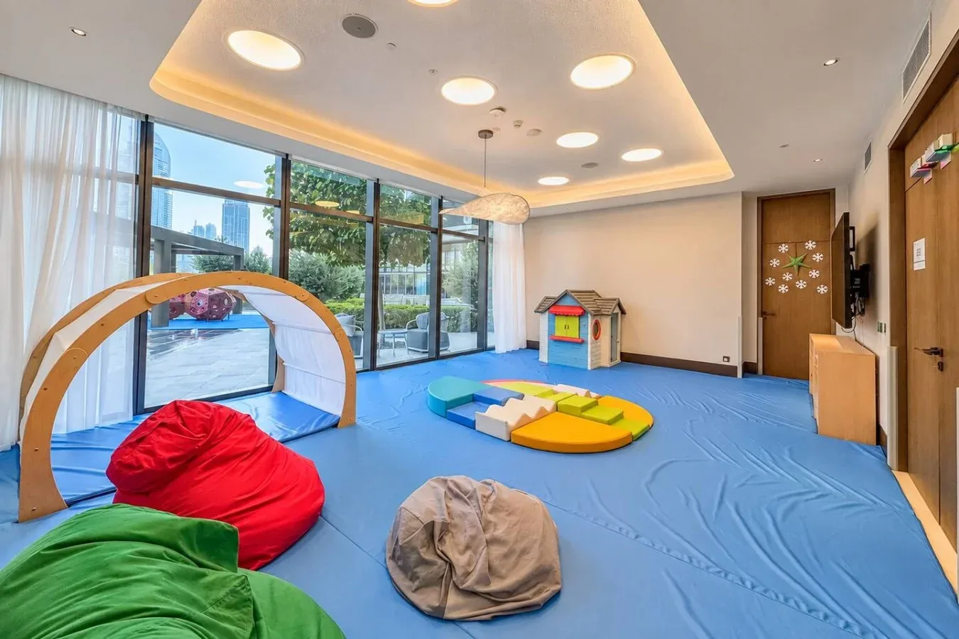 Una luminosa sala de juegos tiene suelo azul, formas acolchadas de colores y grandes ventanas.