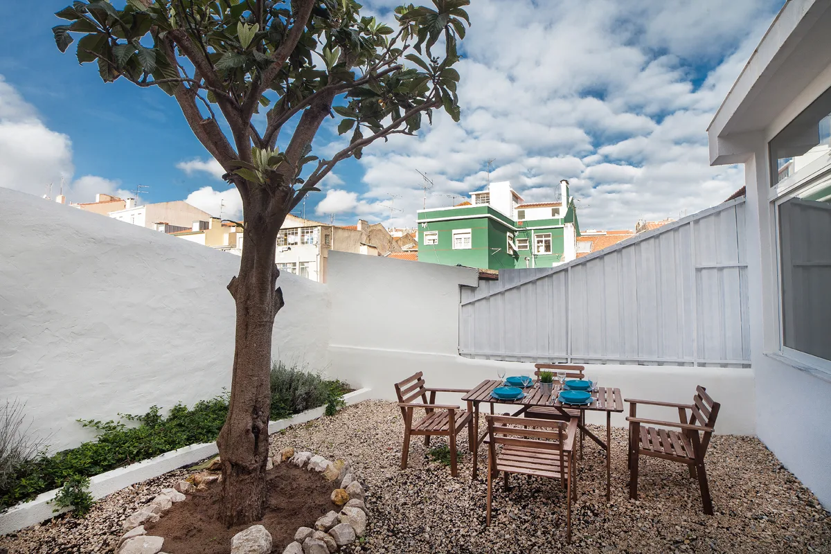 A gravel courtyard with a tree and an outdoor dining table surrounded by wooden seating.