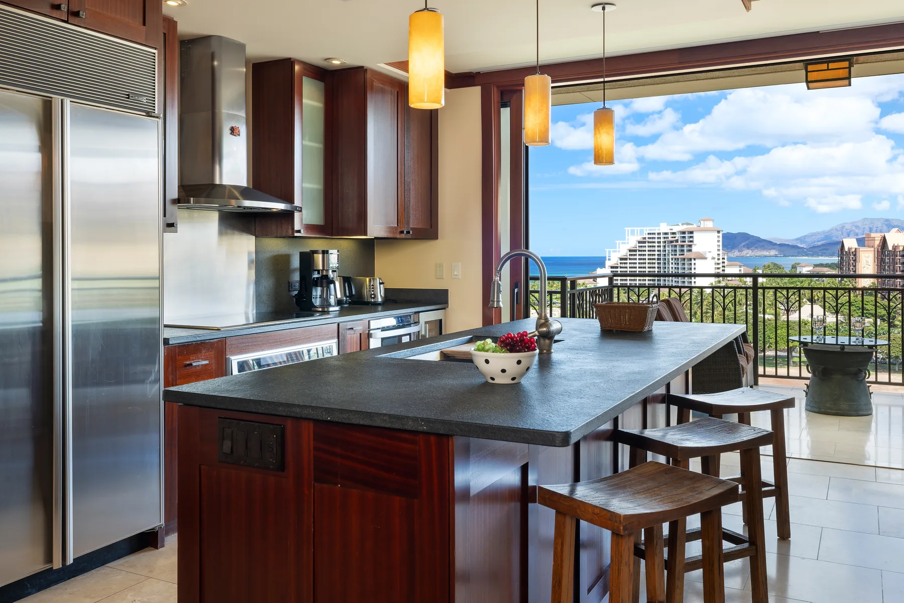 Una isla de cocina con asientos de madera da a un balcón con vistas al océano.