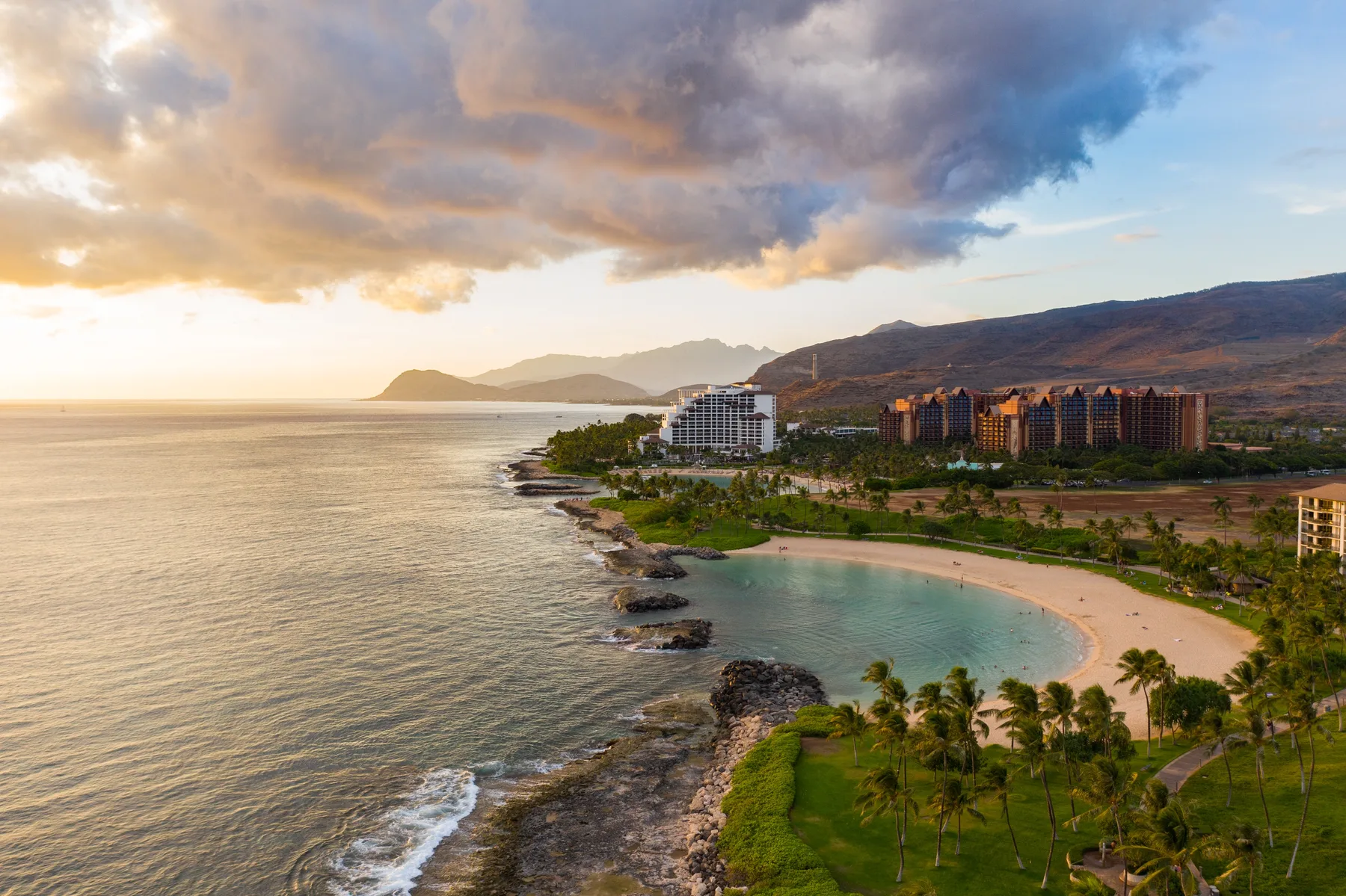 Una playa curva y edificios costeros se encuentran bajo montañas nubladas al atardecer.