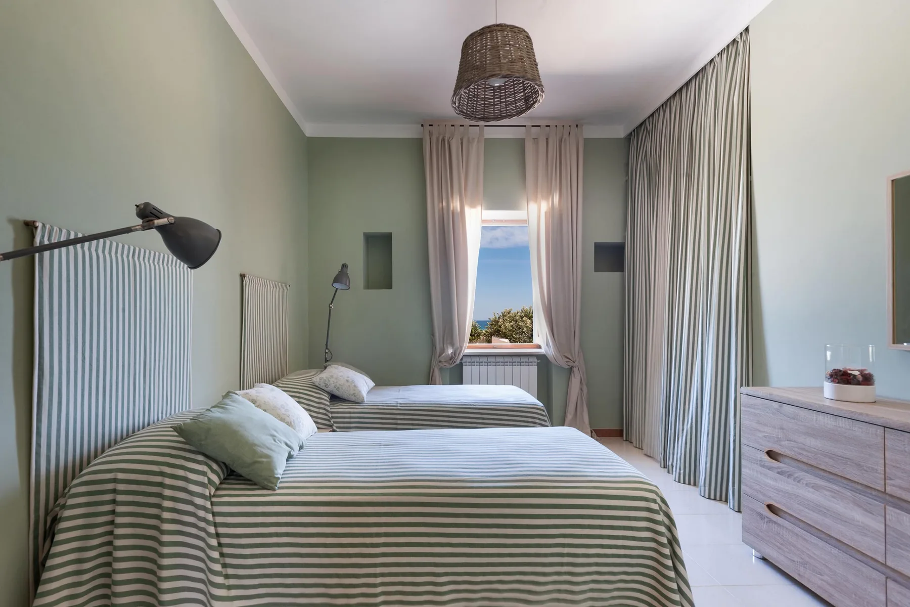 Un dormitorio con ropa de cama a rayas, paredes verde pálido y cortinas alrededor de una ventana.