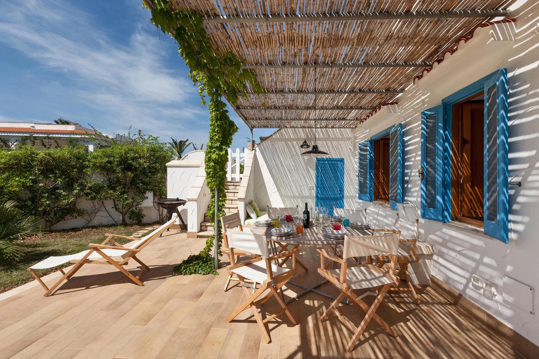 Un patio comedor al aire libre, sombreado, con asientos de madera junto a un edificio blanco.