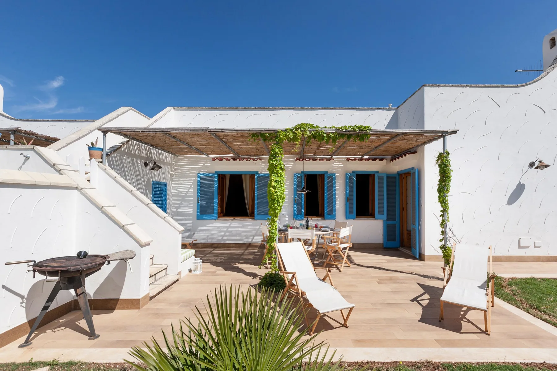 Una terraza soleada con paredes blancas, contraventanas azules y una zona de estar a la sombra.