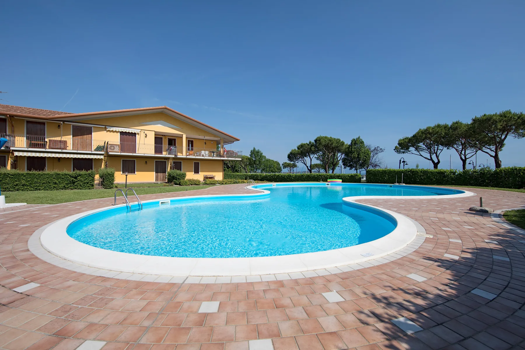 A curved swimming pool sits beside a paved patio and low building.