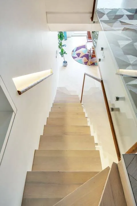 Una escalera de madera desciende hacia una habitación luminosa con una alfombra redonda de colores.