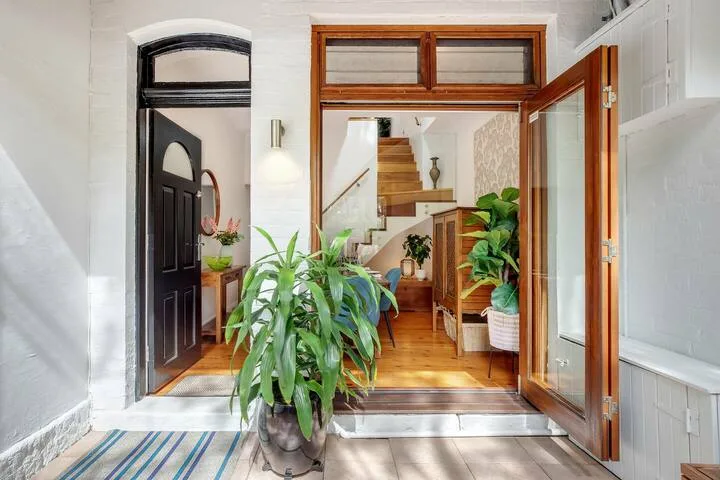 Una entrada abierta da paso a un interior amueblado con escaleras de madera y plantas en macetas.