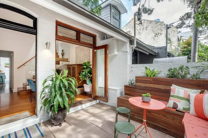 Un patio con asientos de madera, plantas en macetas y una mesa pequeña junto a una puerta abierta.