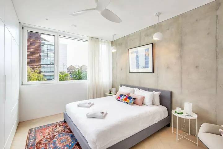 Un dormitorio luminoso con una cama grande junto a ventanas y una pared de aspecto concreto.