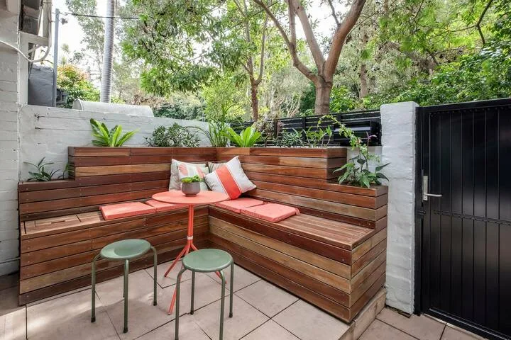 Un patio exterior frondoso tiene asientos de madera empotrados dispuestos en una esquina.