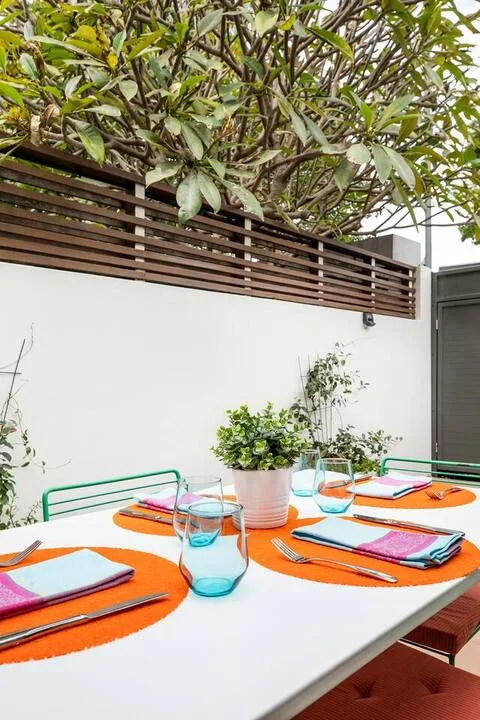 Una mesa de comedor al aire libre está preparada con manteles individuales coloridos y cristalería bajo un árbol frondoso.