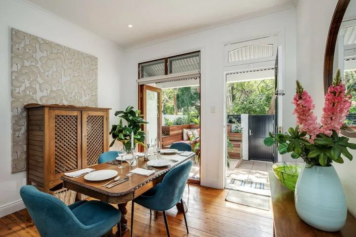 Un comedor luminoso con mesa de madera, asientos azules y una puerta abierta hacia la vegetación.