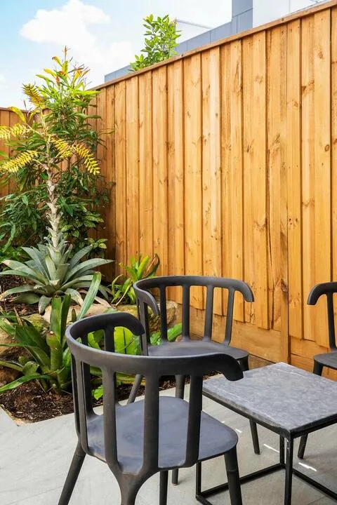 Un patio exterior con asientos junto a una cerca de madera alta y plantas frondosas.