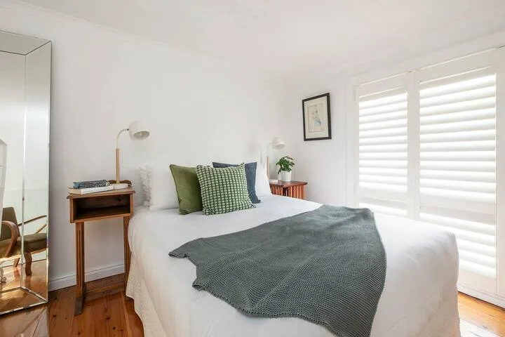 Un dormitorio luminoso con una cama bien tendida y grandes ventanas con persianas.