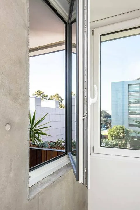 Una ventana luminosa da a una pequeña terraza con plantas y edificios cercanos.