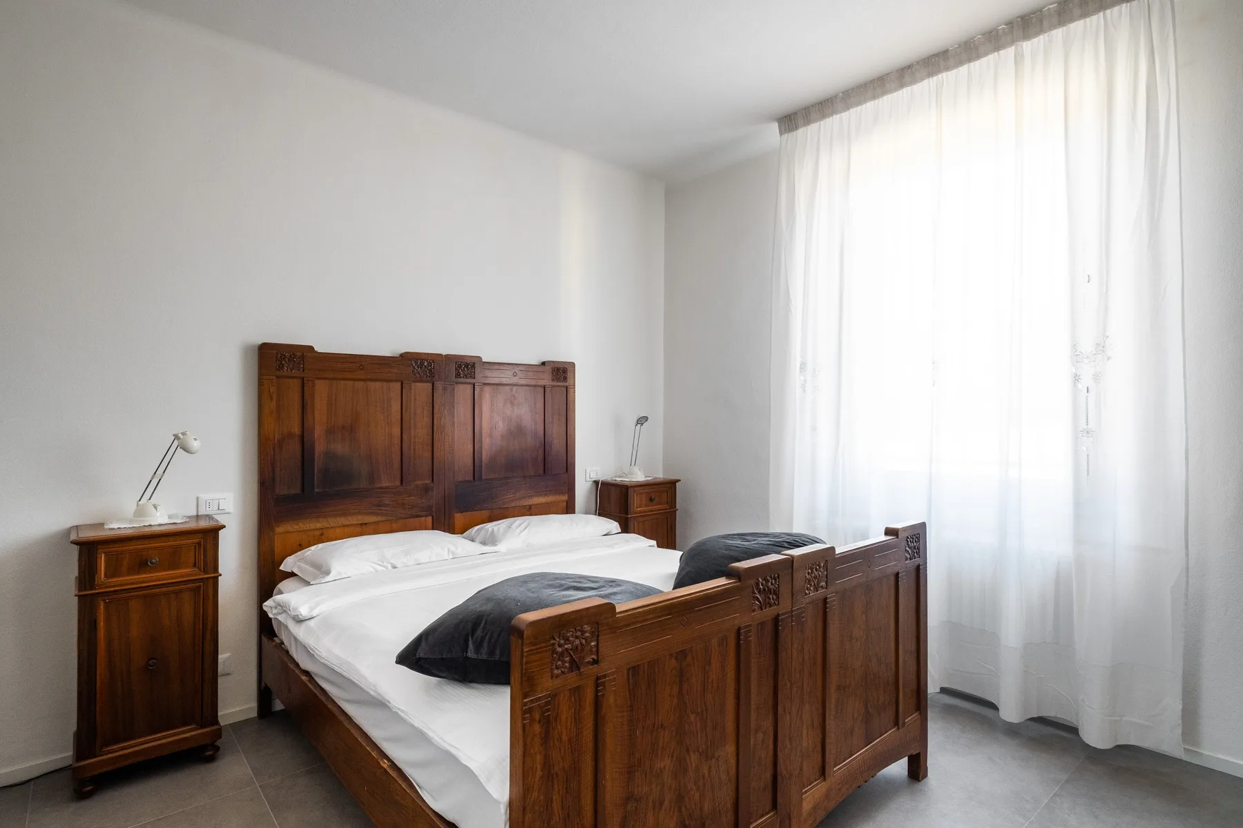 A bedroom with a wooden bed dressed in white bedding beside a bright curtained window.