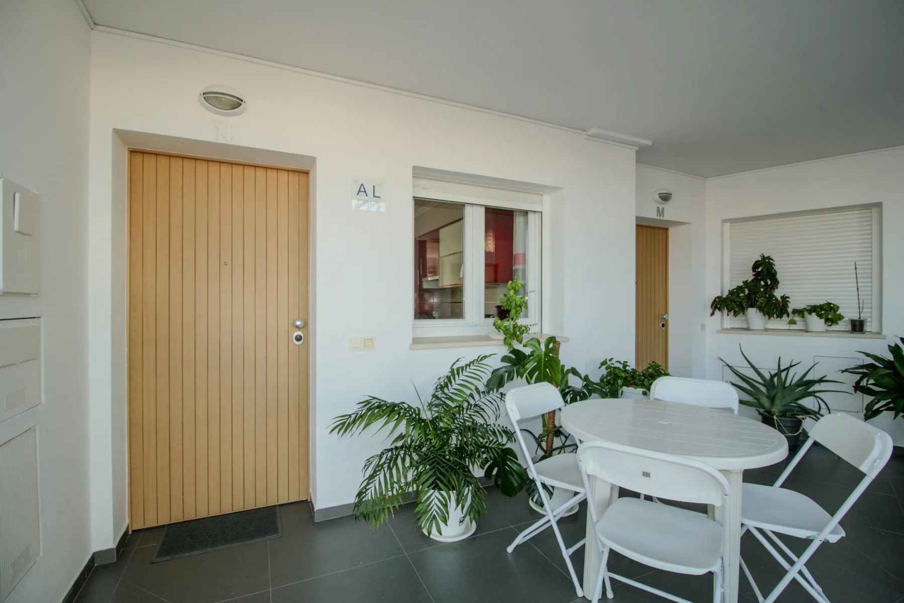 Un patio cubierto y luminoso tiene una mesa redonda, asientos blancos y plantas en macetas.