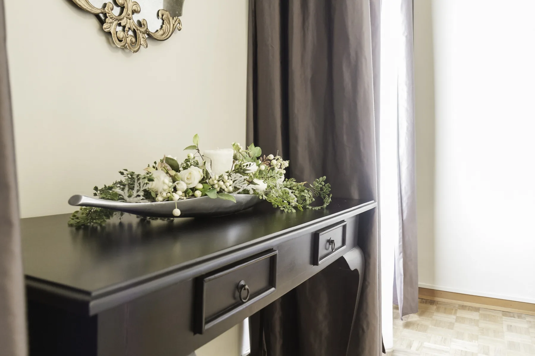 A dark console table with a floral arrangement stands beside a curtained window.