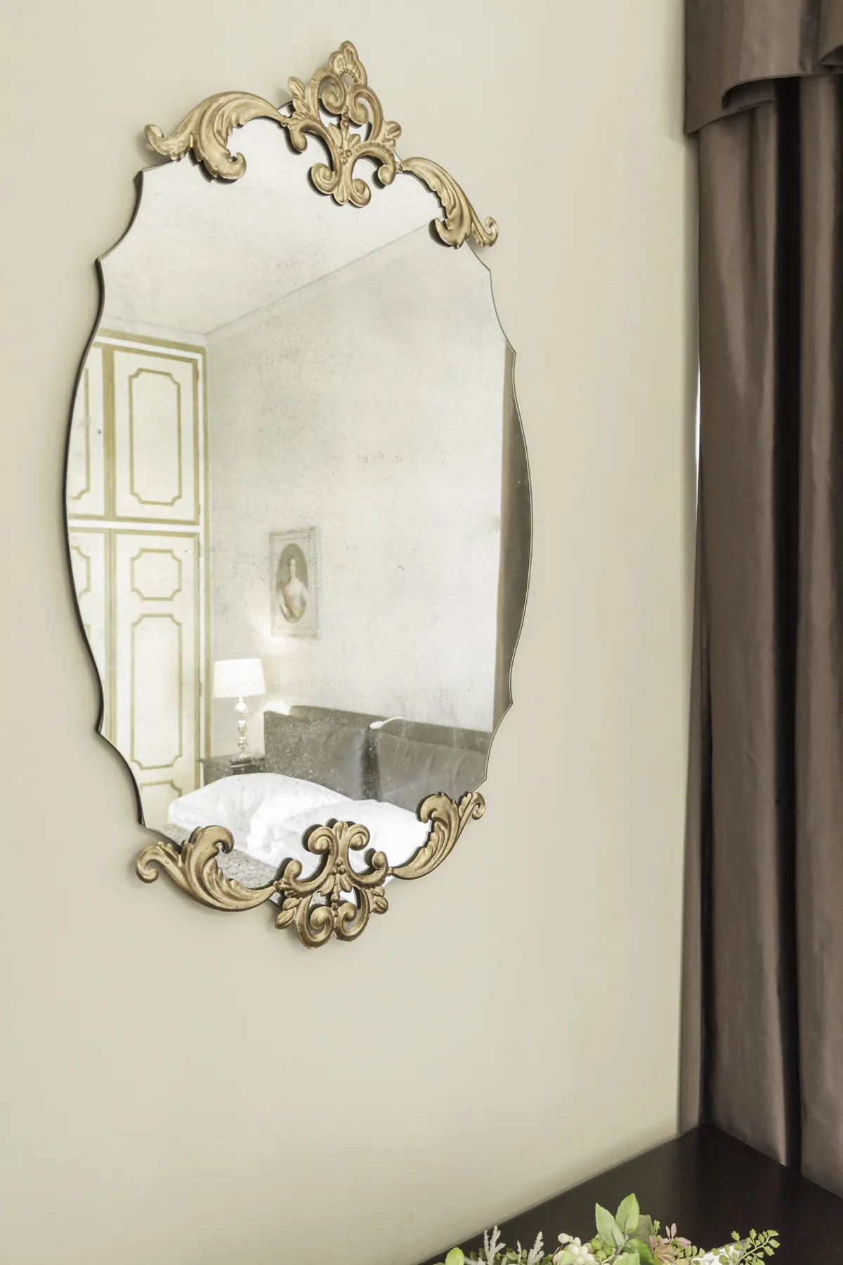 An ornate wall mirror reflects a bedroom with a bed and a bedside lamp.