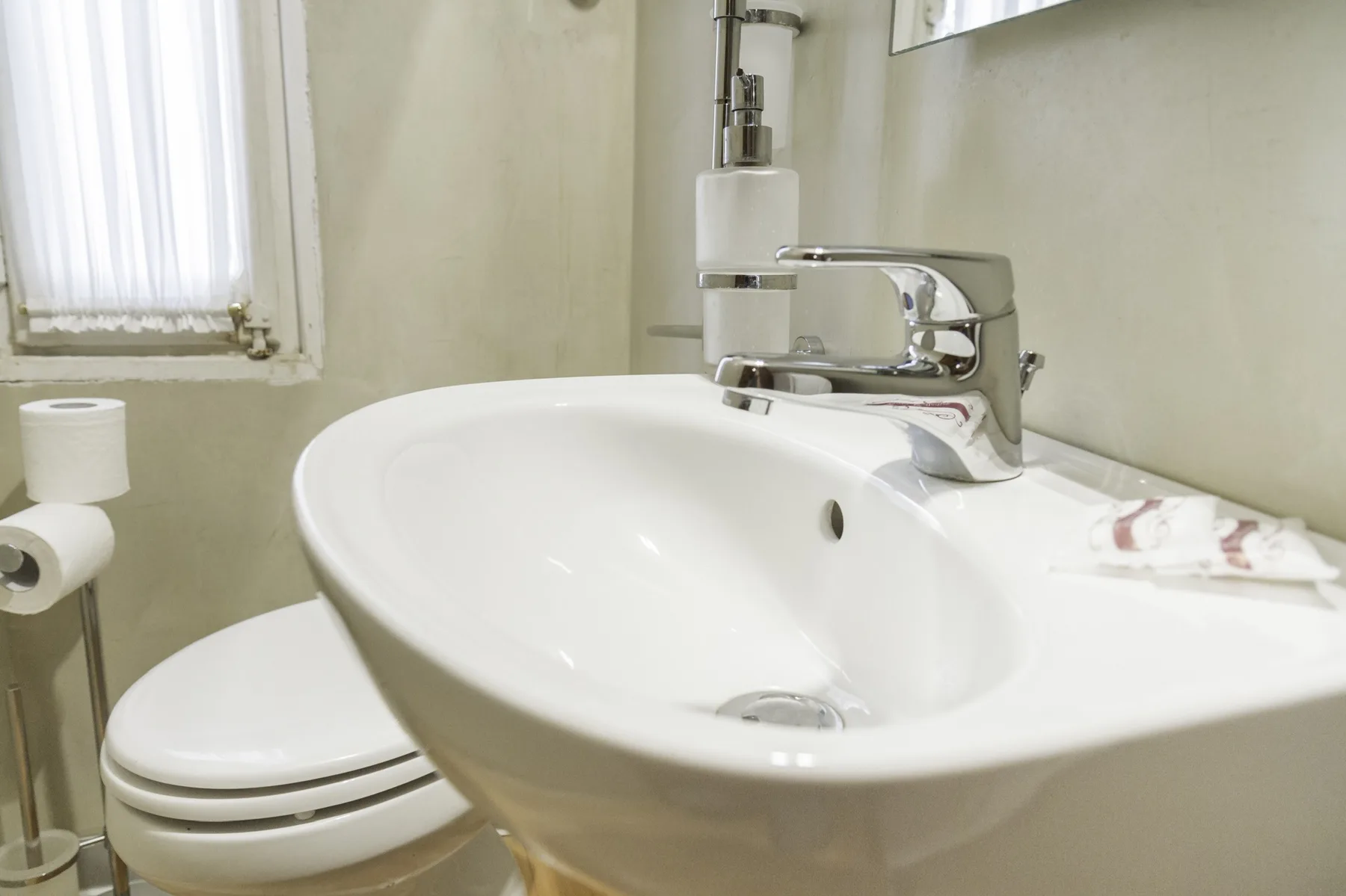 A bathroom sink with a faucet sits beside a toilet and a curtained window.
