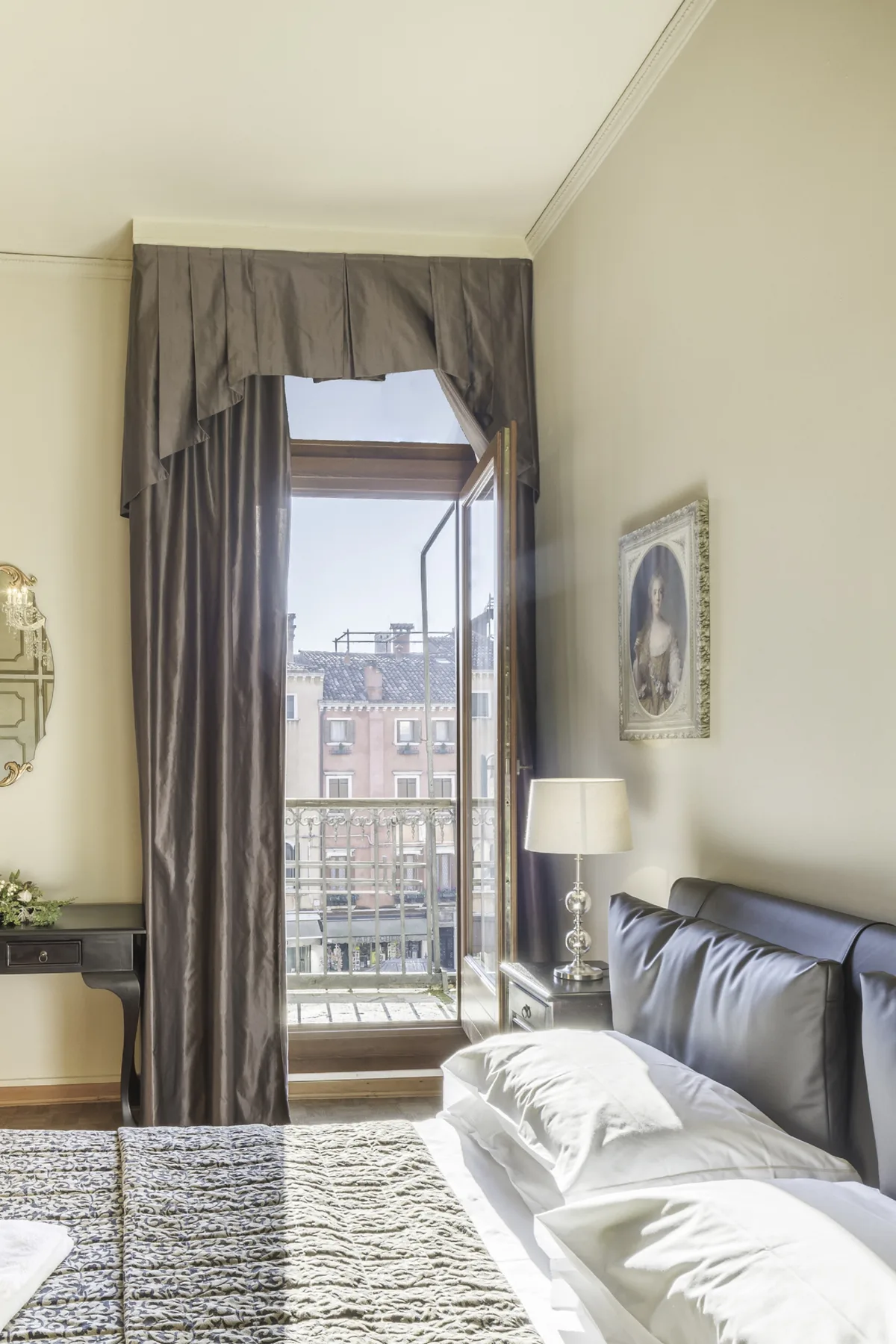 A bedroom with a bed beside an open window overlooking nearby buildings.