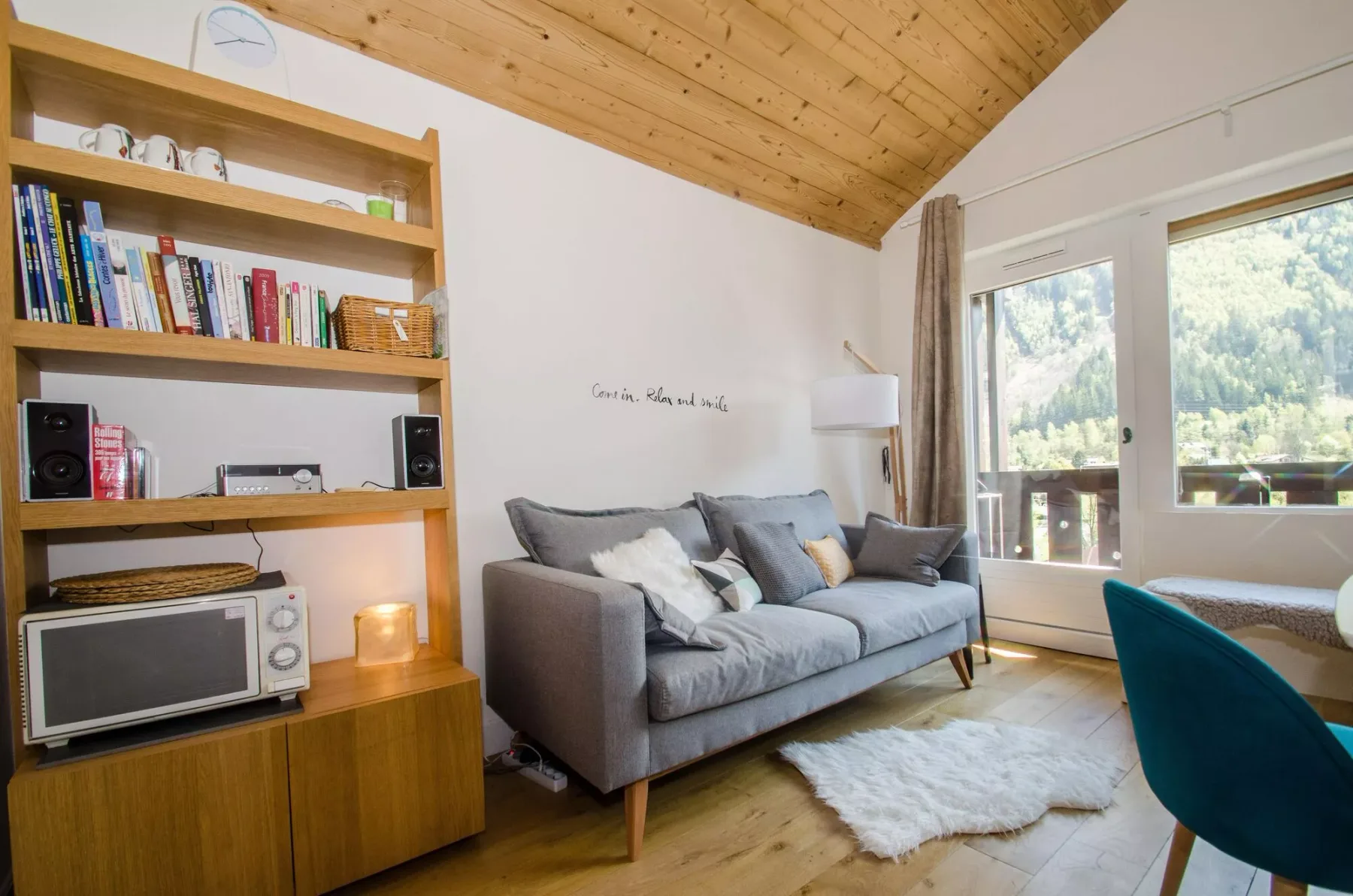 A bright living room with gray seating, wooden shelving, and large windows.