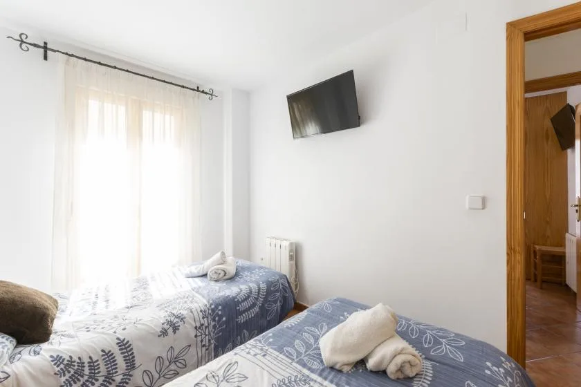 A bright bedroom with two beds, sheer curtains, and a wall-mounted television.