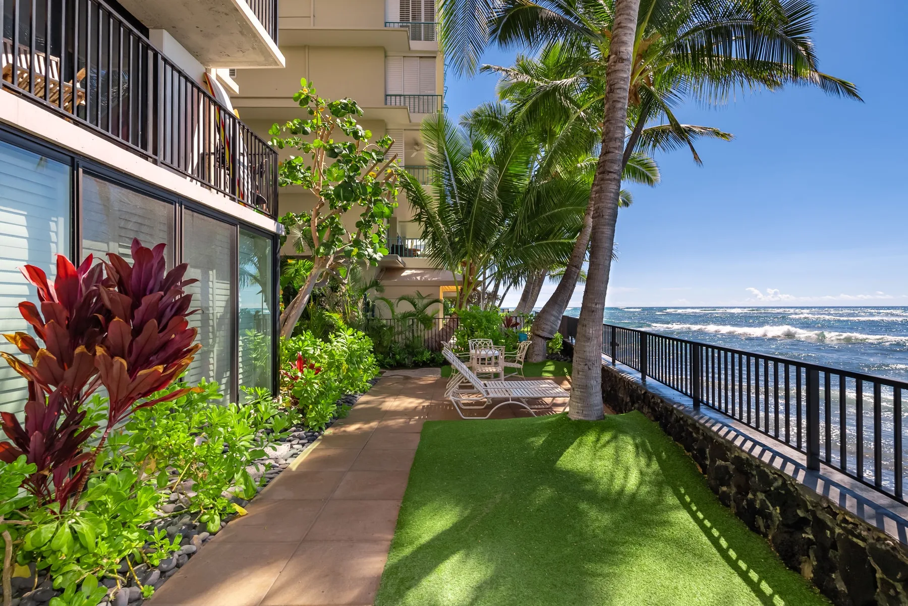 Un patio junto al mar con césped verde, plantas tropicales y vistas al océano.