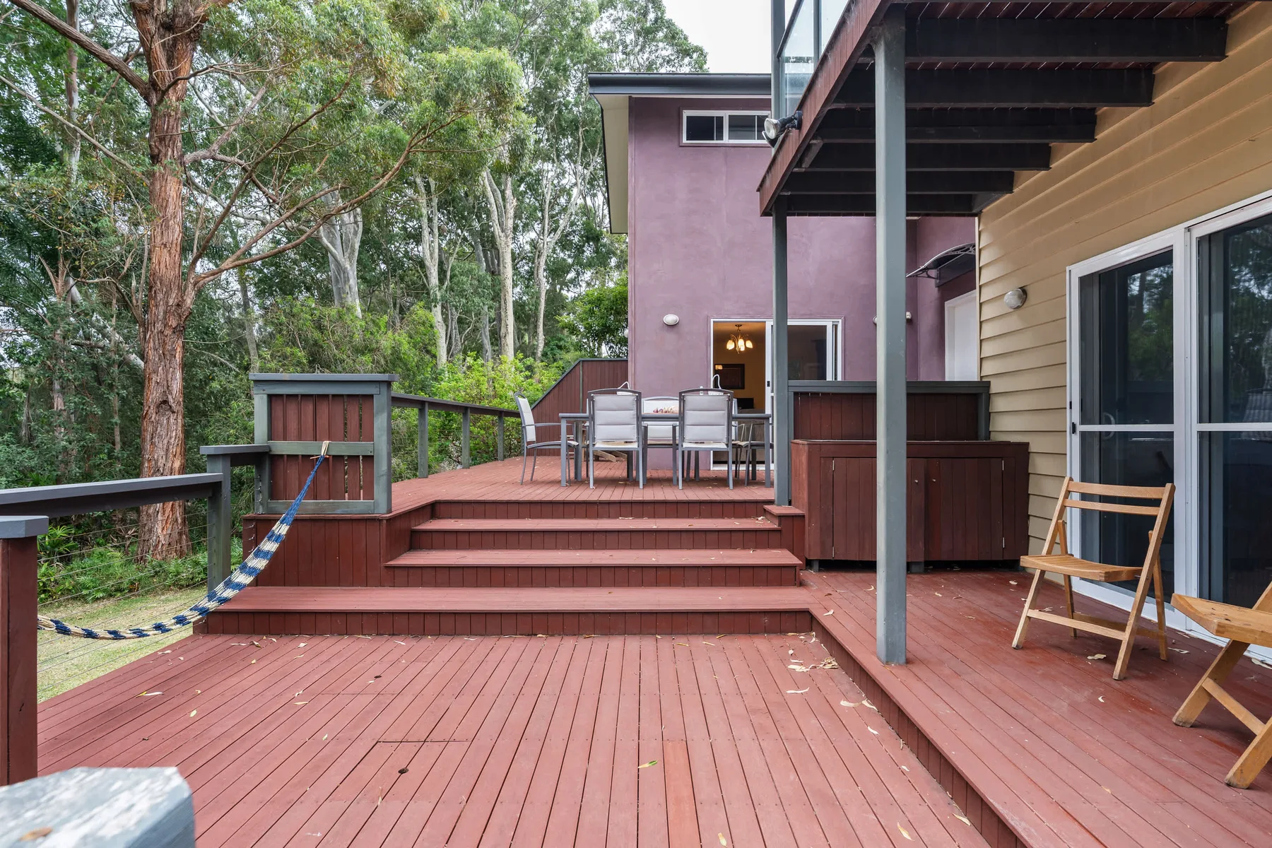 Una terraza exterior de madera con asientos y un jardín arbolado al fondo.