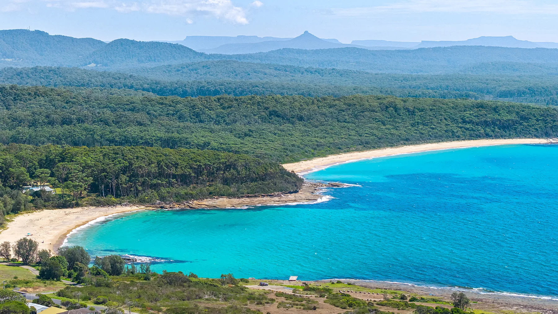Una playa de arena curva bordea una bahía turquesa y colinas boscosas.