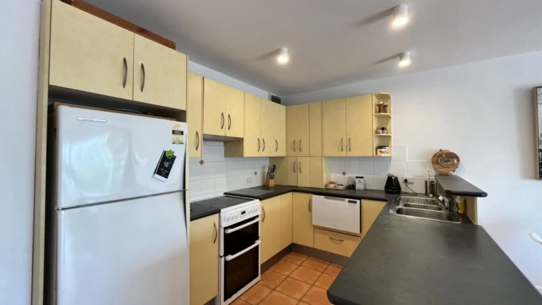 A kitchen with light cabinets, a refrigerator, and dark countertops.