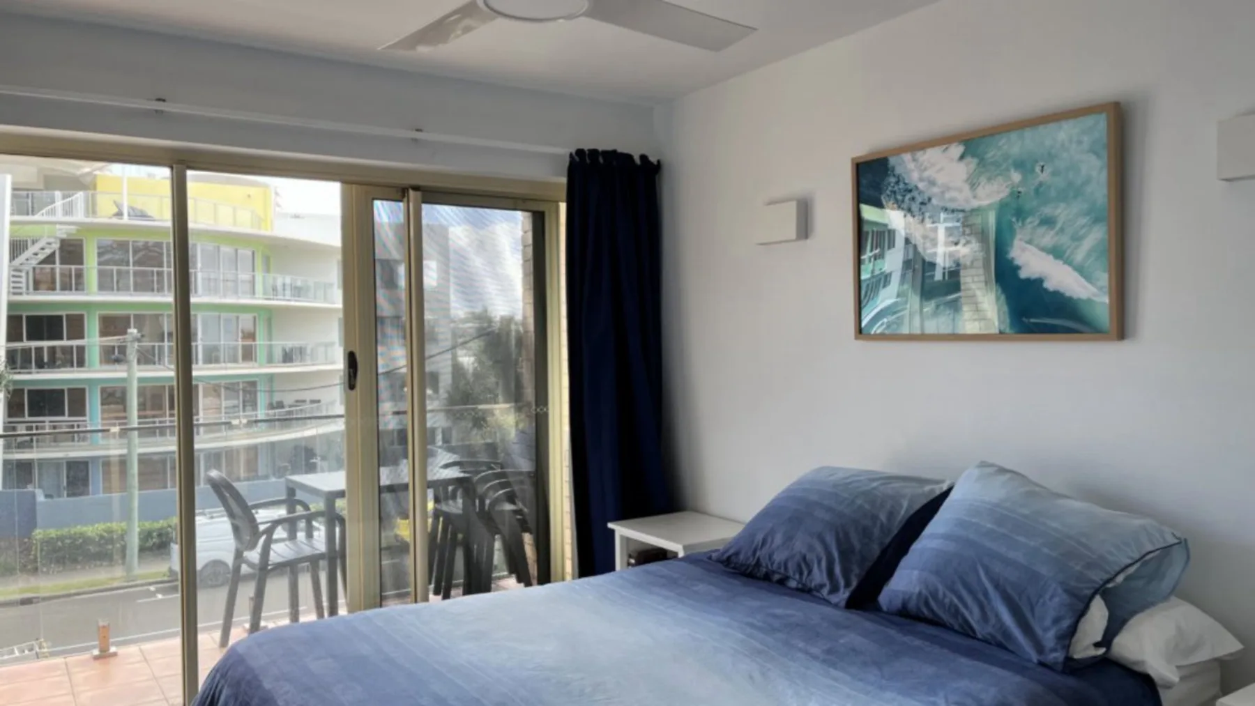 A bedroom with a blue bed beside sliding glass doors opening onto a balcony.