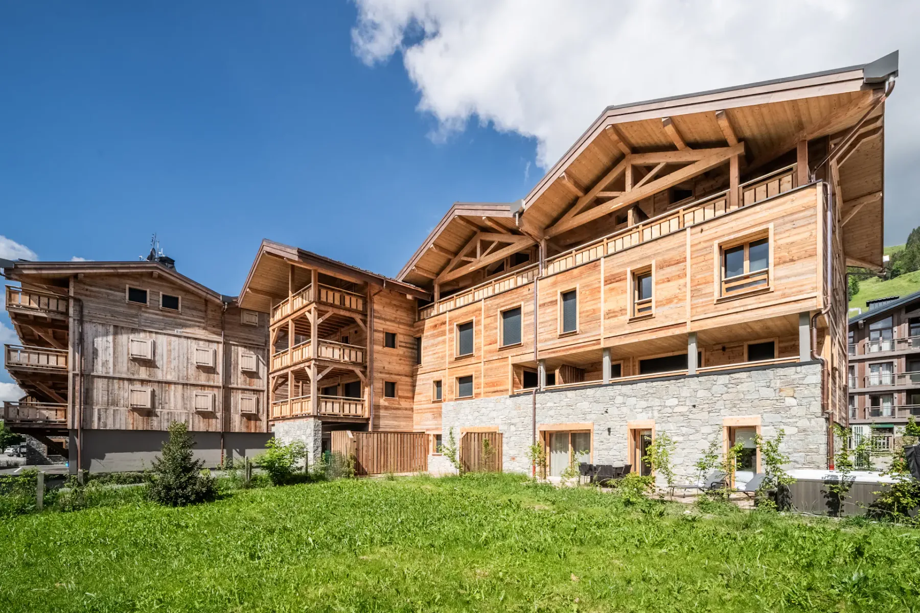 A cluster of wooden chalet-style buildings with stone foundations and balconies, set against a blue sky with patches of clouds and a lawn in the foreground.