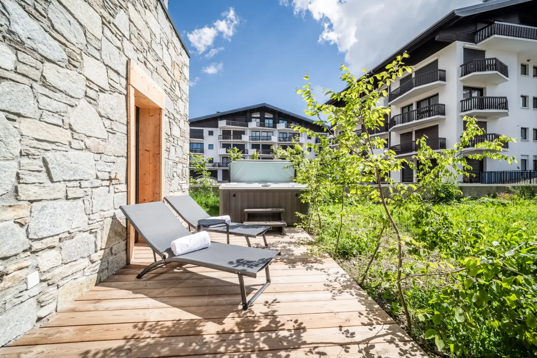 A wooden outdoor terrace has lounge seating beside a hot tub, with green plants and buildings beyond.